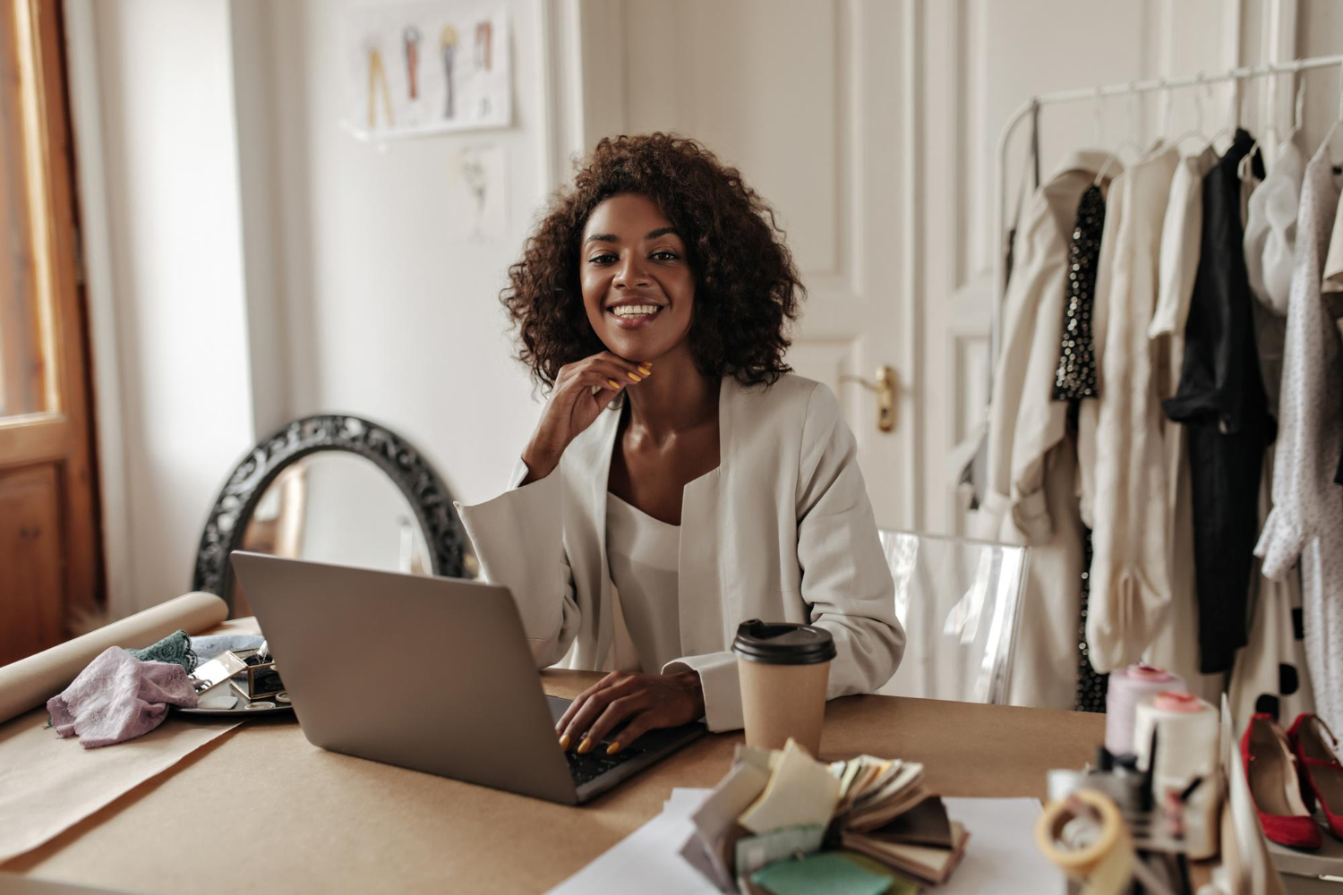 Mulher negra vestida com blazer, sentada à mesa com computador e copo de café olha com sorriso simpático