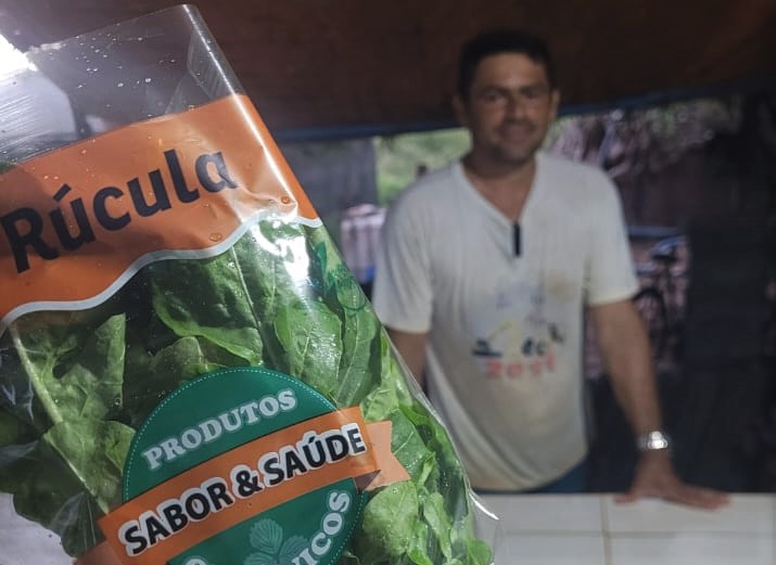 Pacote de rúcula aparece em primeiro plano com o produtor Tiago ao fundo de camisa branca